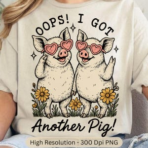 May include: Beige t-shirt featuring a graphic of two smiling pigs wearing heart-shaped sunglasses. The text above reads "OOPS! I GOT" and below "Another Pig!". Yellow flowers and green grass are at the bottom of the image.