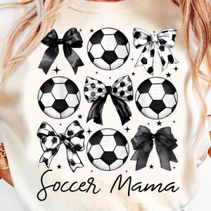 Puede incluir: Camiseta blanca con un dise&ntilde;o gr&aacute;fico en blanco y negro de balones de f&uacute;tbol y lazos. El texto "Soccer Mama" est&aacute; impreso en la camiseta.