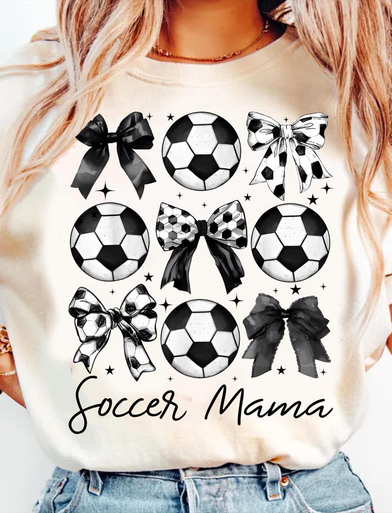 Puede incluir: Camiseta blanca con un dise&ntilde;o gr&aacute;fico en blanco y negro de balones de f&uacute;tbol y lazos. El texto "Soccer Mama" est&aacute; impreso en la camiseta.