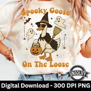 May include: Cream-colored t-shirt featuring a Halloween-themed graphic. The design includes a goose wearing a witch hat and sunglasses, holding a broom and a pumpkin basket. Text reads "Spooky Goose On The Loose."