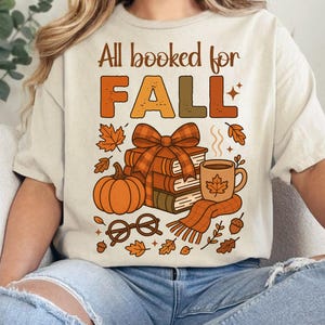 May include: Beige t-shirt with the text "All booked for FALL" in orange and brown. The graphic includes a pumpkin, books, a coffee mug, and fall leaves. A cozy autumn-themed design.