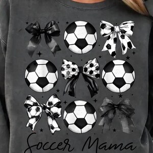 Puede incluir: Sudadera gris con un dise&ntilde;o de bal&oacute;n de f&uacute;tbol y lazo en blanco y negro. El texto "Soccer Mama" est&aacute; impreso en la parte inferior de la sudadera.