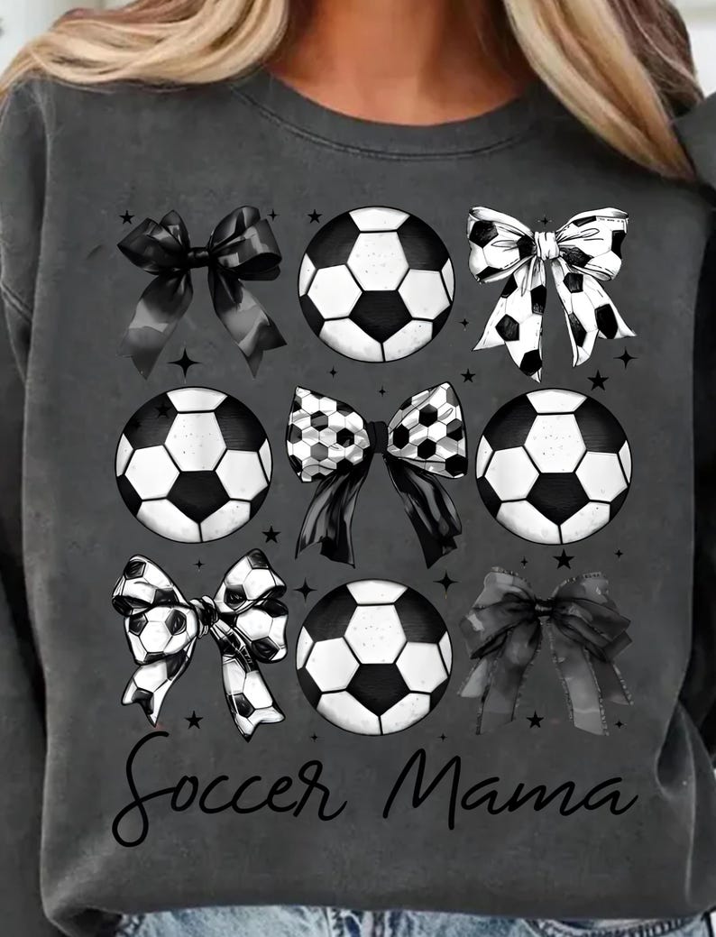 Puede incluir: Sudadera gris con un dise&ntilde;o de bal&oacute;n de f&uacute;tbol y lazo en blanco y negro. El texto "Soccer Mama" est&aacute; impreso en la parte inferior de la sudadera.