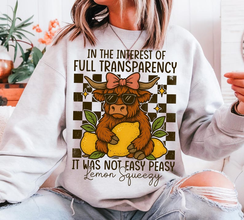 May include: A white sweatshirt with a graphic of a brown and white cow holding a lemon. The text on the sweatshirt reads "In the interest of full transparency it was not easy peasy lemon squeezy."