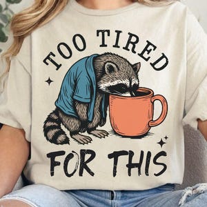 Op de afbeelding: Beige t-shirt met een illustratie van een wasbeer, gekleed in een blauw jasje, drinkend uit een oranje mok. De tekst "TOO TIRED FOR THIS" staat boven en onder de wasbeer-afbeelding. Het ontwerp bevat kleine steraccenten.