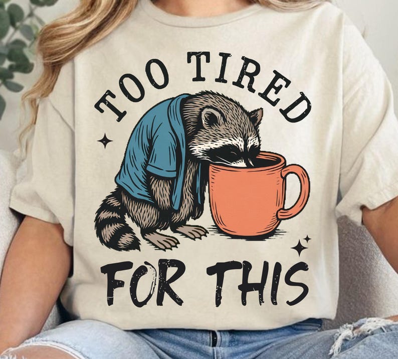 Op de afbeelding: Beige t-shirt met een illustratie van een wasbeer, gekleed in een blauw jasje, drinkend uit een oranje mok. De tekst "TOO TIRED FOR THIS" staat boven en onder de wasbeer-afbeelding. Het ontwerp bevat kleine steraccenten.