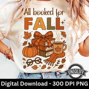 May include: Cream-colored t-shirt with a fall-themed graphic. The design includes the text "All booked for FALL" in orange and brown, along with a pumpkin, books, a coffee mug, and fall leaves. A cozy autumn design.