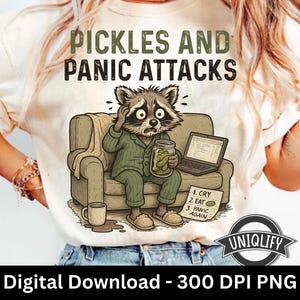 May include: A cream t-shirt featuring the text "PICKLES AND PANIC ATTACKS" and a cartoon raccoon in pyjamas. The raccoon, holding a pickle jar, sits on a sofa with a laptop and a list reading "1. CRY, 2. EAT, 3. PANIC AGAIN."