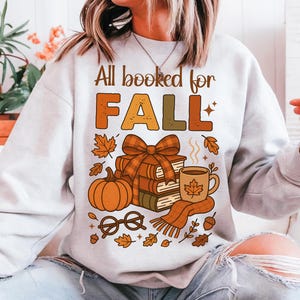 May include: A light gray sweatshirt with the text "All booked for FALL" in brown and orange. The graphic includes a stack of books, a pumpkin, a coffee mug, and fall leaves.