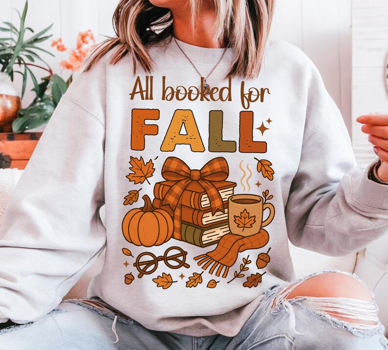 May include: A light gray sweatshirt with the text "All booked for FALL" in brown and orange. The graphic includes a stack of books, a pumpkin, a coffee mug, and fall leaves.