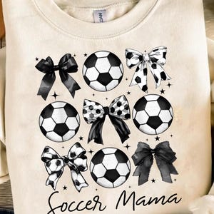 Puede incluir: Una sudadera de color crema con el texto "Soccer Mama" en letras cursivas negras. El dise&ntilde;o presenta balones de f&uacute;tbol y lazos en blanco y negro.