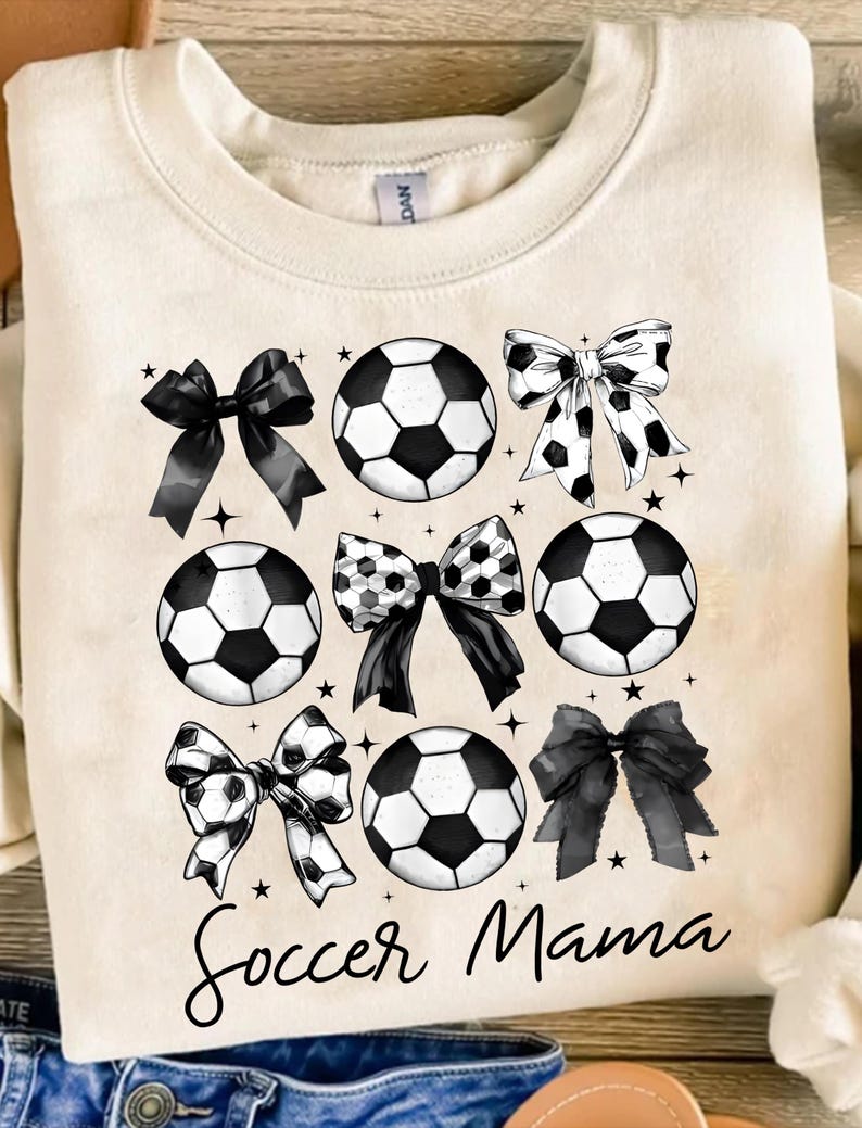 Puede incluir: Una sudadera de color crema con el texto "Soccer Mama" en letras cursivas negras. El dise&ntilde;o presenta balones de f&uacute;tbol y lazos en blanco y negro.