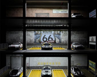 Op de afbeelding: Een parkeergarage op schaal met meerdere verdiepingen en zes miniatuur sportwagens in geel gemarkeerde vakken. De structuur bevat een Route 66-bord op de achterwand, structurele steunpilaren en witte richtingspijlen op de vloer.
