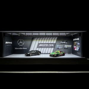 May include: A black Mercedes-Benz AMG car and a green and black AMG race car are displayed in a showroom setting. The background features a large banner with the Mercedes-Benz logo and the words "Mercedes-Benz" and "AMG. Drive you further." The banner also features a smaller banner with the words "AMG. Once you have nothing, you have everything."
