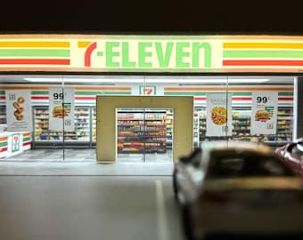 May include: A miniature 7-Eleven store model featuring a bright illuminated sign with green, orange, and red stripes. The storefront has glass windows displaying posters with food graphics and the number 99, with a blurred car in the foreground.
