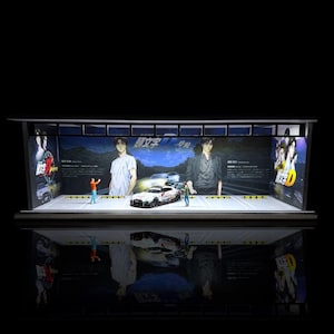 May include: A white and blue model car with a black and red stripe on the side. The car is on a display stand with a backdrop of a poster featuring anime characters and the text "Initial D".