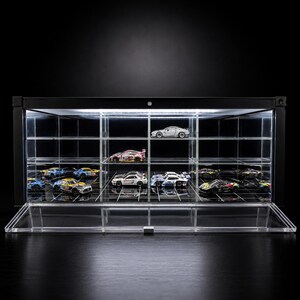 May include: A black rectangular display case with clear acrylic shelves and a front-opening door. The interior features integrated LED lighting illuminating several miniature toy race cars arranged on the tiered shelving system.