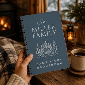 Puede incluir: Un cuaderno de espiral azul con el texto "The MILLER FAMILY" y "GAME NIGHT SCOREBOOK" impreso en blanco. La portada presenta una ilustración de líneas blancas de una cabaña entre pinos bajo una pequeña estrella.