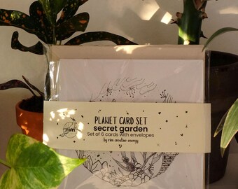 May include: A set of six white greeting cards with black line art illustrations, packaged with envelopes. The packaging includes a beige band with the text "PLANET CARD SET secret garden" and the artist's name. The cards are surrounded by green plants.