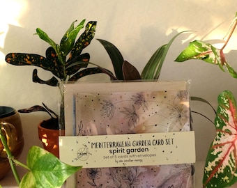 May include: A clear plastic-wrapped card set with a floral design and the text "MEDITERRANEAN GARDEN CARD SET spirit garden". The set includes 5 cards with envelopes. The background features green plants and small stones.