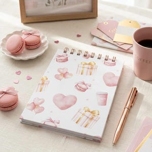 May include: A spiral-bound notebook with a white cover featuring pink watercolor illustrations of hearts, gifts, macarons, and coffee cups. A rose gold pen, pink macarons, and a pink coffee mug are also visible. A framed floral print and gift tags complete the scene.