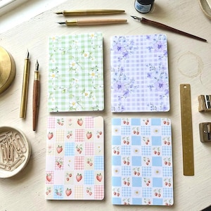 May include: Four notebooks with floral and fruit patterns are arranged on a white surface. The notebooks feature designs of green gingham with white flowers, purple floral patterns, pink and blue patchwork with strawberries, and blue patchwork with strawberries and daisies. Gold pens and ink bottles are also visible.