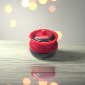 May include: A small, decorative jar with a lid. The jar is primarily red with a black band around the middle and a green base. The lid is also red with a small knob on top. The jar is sitting on a light-colored, textured surface.