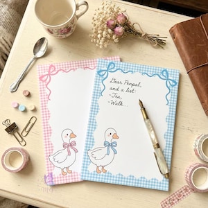 May include: Two decorative notepads with a gingham border, one pink and one blue, featuring a cartoon goose with a bow. The blue notepad has the text "Dear Penpal, and a list: -Tea, -Walk...". A vintage pen, spoon, and washi tape are also visible.