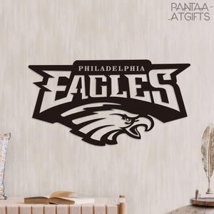 以下が含まれることがあります： フィラデルフィア・イーグルスのロゴとチーム名が描かれた黒い金属製のサイン。明るい色の壁に取り付けられています。チーム名は、ワシのグラフィックの上に表示されています。