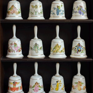 Puede incluir: Doce campanas de porcelana blanca con detalles dorados, cada una con un diseño de hada diferente. Las campanas se exhiben en una estantería de madera marrón oscuro con tres niveles. Las ilustraciones de hadas incluyen varios colores y detalles florales.
