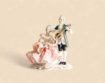 May include: A porcelain figurine depicting a man playing a lute and a woman seated with a fan. The woman wears a pink and white dress, while the man is in a teal coat and black shorts. The base is white with gold accents.