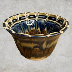 May include: A ceramic bowl featuring a fluted, scalloped rim with a cream-coloured edge. The interior and exterior display a reactive glaze finish with deep blue, dark brown, and tan tones that blend in a flowing, drip-like pattern across the surface.