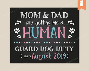 May include: A black chalkboard sign with white text announcing that Mom and Dad are getting a human. The text is in pink and blue, with paw prints and bone graphics. The sign also says "Guard Dog Duty starts August 2019".