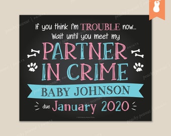 May include: A black chalkboard sign with white text announcing the arrival of a new baby. The text reads "If you think I'm trouble now... Wait until you meet my partner in crime. Baby Johnson due January 2020." The sign is decorated with paw prints and bone graphics.