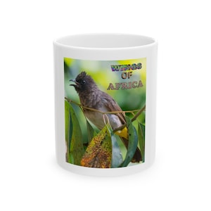 Puede incluir: Taza de cerámica blanca con una imagen colorida de un pájaro posado en una rama con hojas verdes. La taza tiene el texto "WINGS OF AFRICA" en letras coloridas. Un artículo único y llamativo, ideal para té, café o cualquier bebida.