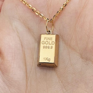 May include: A gold chain necklace with a gold pendant shaped like a gold bar. The pendant has the words "FINE GOLD 999.9 1Kg" engraved on it.
