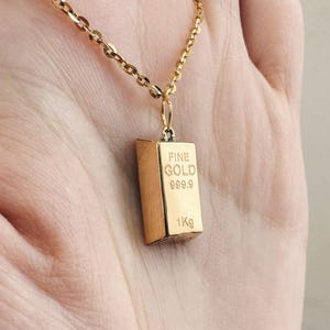 May include: A gold chain necklace with a gold pendant shaped like a gold bar. The pendant has the words "FINE GOLD 999.9 1Kg" engraved on it.