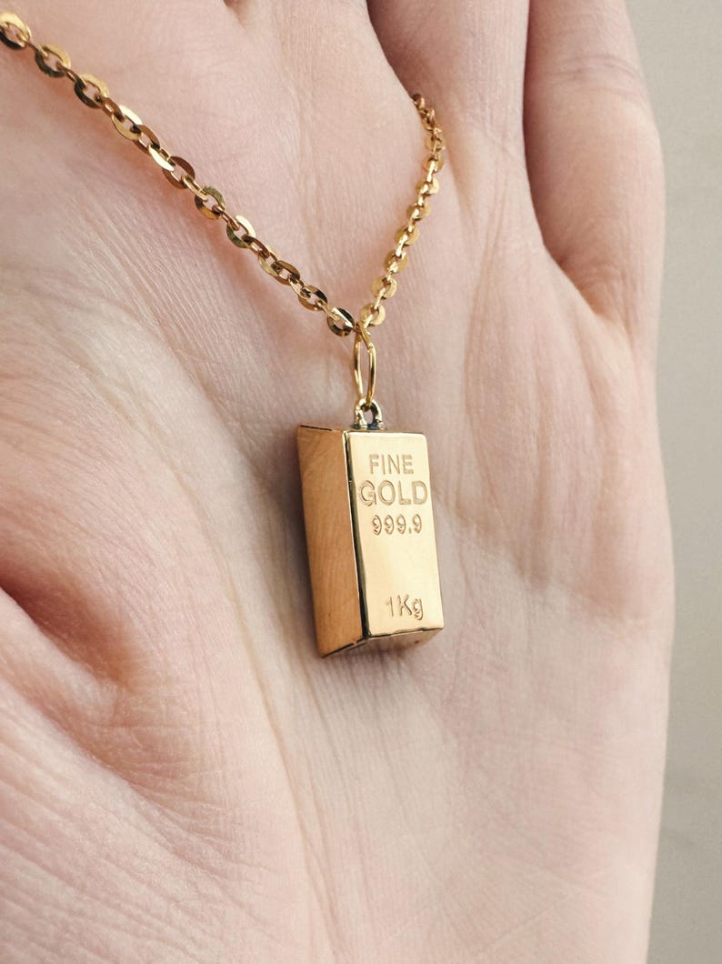 May include: A gold chain necklace with a gold pendant shaped like a gold bar. The pendant has the words "FINE GOLD 999.9 1Kg" engraved on it.