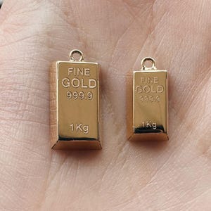 May include: Two gold-colored charms shaped like gold bars. Each charm has the words "FINE GOLD 999.9 1Kg" engraved on it.