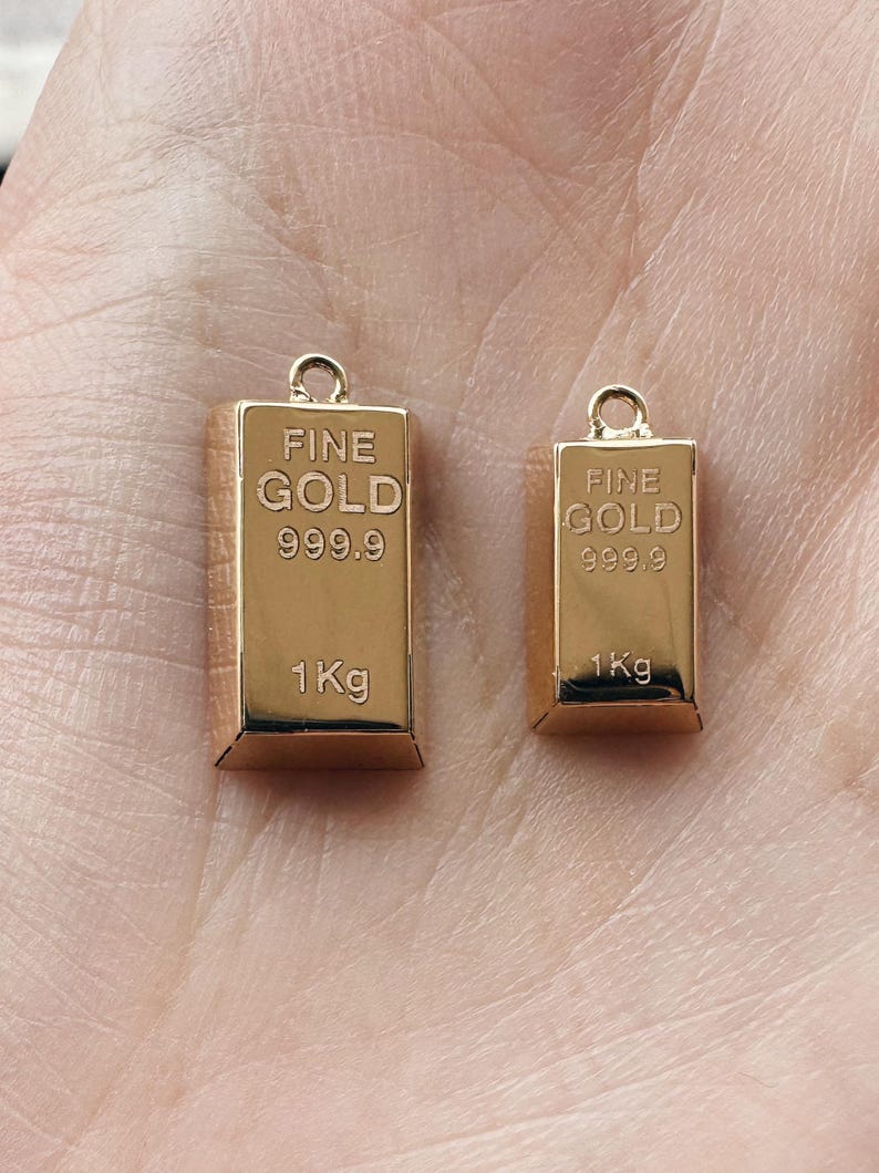 May include: Two gold-colored charms shaped like gold bars. Each charm has the words "FINE GOLD 999.9 1Kg" engraved on it.