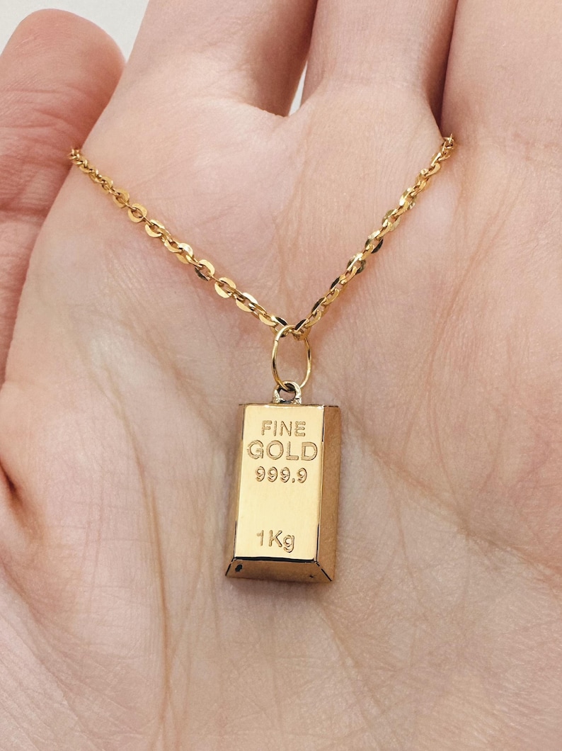 May include: A gold chain necklace with a gold pendant shaped like a gold bar. The pendant has the words "FINE GOLD 999.9 1Kg" engraved on it.
