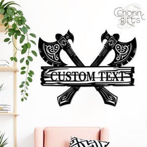 May include: Black metal wall art featuring two crossed axes with ornate detailing. A wooden plank with the text "CUSTOM TEXT" is centered between the axes. The design is suitable for home decor.