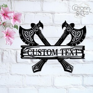 May include: Black metal wall art featuring two crossed axes with ornate detailing. A wooden-style banner across the axes reads "CUSTOM TEXT". The axes are set against a white brick wall, with pink flowers in the upper left corner.