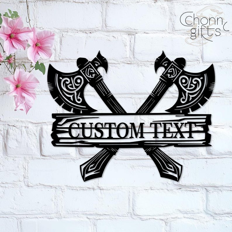 May include: Black metal wall art featuring two crossed axes with ornate detailing. A wooden-style banner across the axes reads "CUSTOM TEXT". The axes are set against a white brick wall, with pink flowers in the upper left corner.