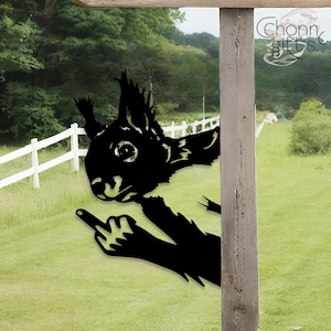 May include: Black metal squirrel silhouette with a raised middle finger, set against a green lawn and white fence. The squirrel is partially obscured by a wooden post. The image has the text "Chonn gifts" in the upper right corner.