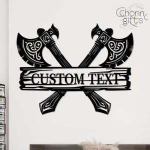 May include: Black metal wall art featuring two crossed axes with ornate detailing. A wooden plank in the center displays the text "CUSTOM TEXT". The design includes decorative elements and the brand name "Chonn gifts".