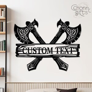 May include: Black metal wall art featuring two crossed axes with ornate detailing. A horizontal plank in the center displays the words "CUSTOM TEXT". The axes have decorative handles and blades. The overall design is a rustic, Viking-inspired theme.