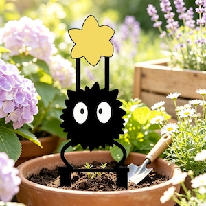 May include: A black metal plant trellis featuring a spiky, circular creature with two large white eyes, topped with a yellow star-shaped accent. The structure has two prongs at the base for insertion into soil, standing upright in a terracotta pot.