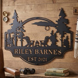 May include: Black metal wall art featuring a camping scene with trees, Adirondack chairs, and a campfire. The sign reads "RILEY BARNES EST 2021". Decorative metal art for home decor.