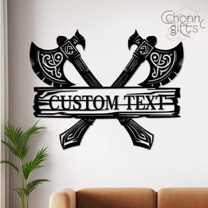 May include: Black metal wall art featuring two crossed axes with ornate detailing. A horizontal plank in the center displays the text "CUSTOM TEXT". The axes have decorative patterns and are set against a white background.